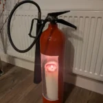 Front view of upcycled fire extinguisher candle holder #3 with a lit pillar candle, looped black hose and discharge horn.