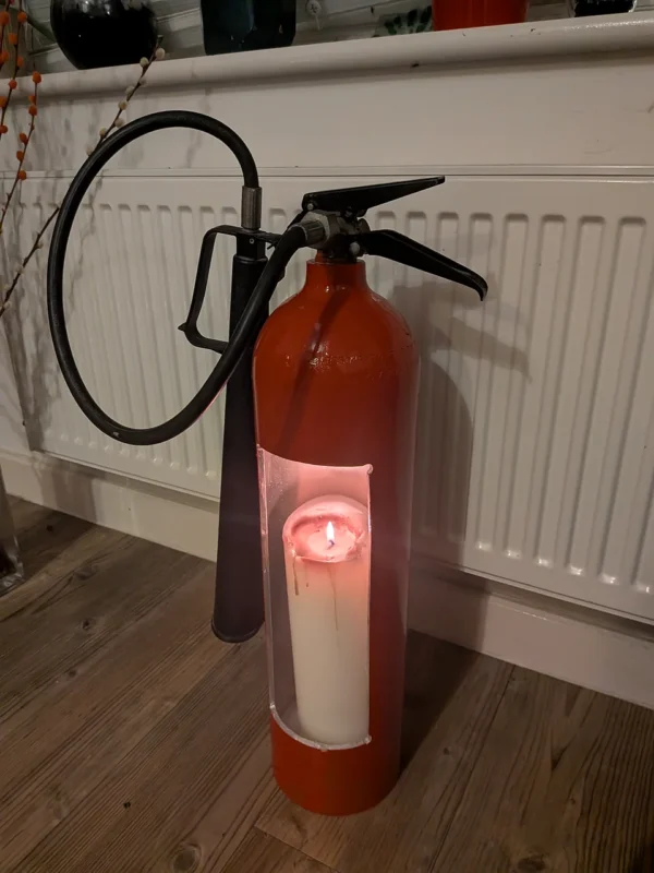 Front view of upcycled fire extinguisher candle holder #3 with a lit pillar candle, looped black hose and discharge horn.