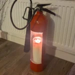 Red upcycled fire extinguisher candle holder #3 displayed on a wooden floor with candlelight visible through the opening.