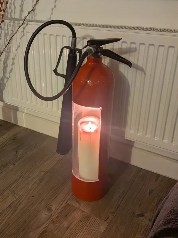 Red upcycled fire extinguisher candle holder #3 displayed on a wooden floor with candlelight visible through the opening.