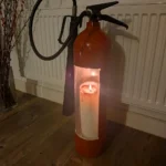Low-light view of red upcycled fire extinguisher candle holder #3 with a glowing pillar candle inside.