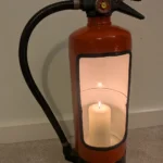 Front view of upcycled fire extinguisher candle holder #9 with a lit pillar candle, original black hose and pressure gauge.