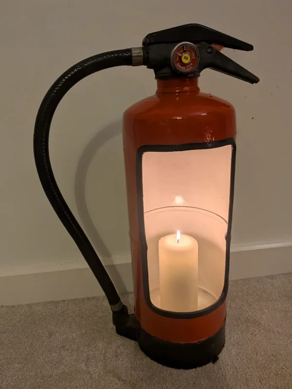 Front view of upcycled fire extinguisher candle holder #9 with a lit pillar candle, original black hose and pressure gauge.